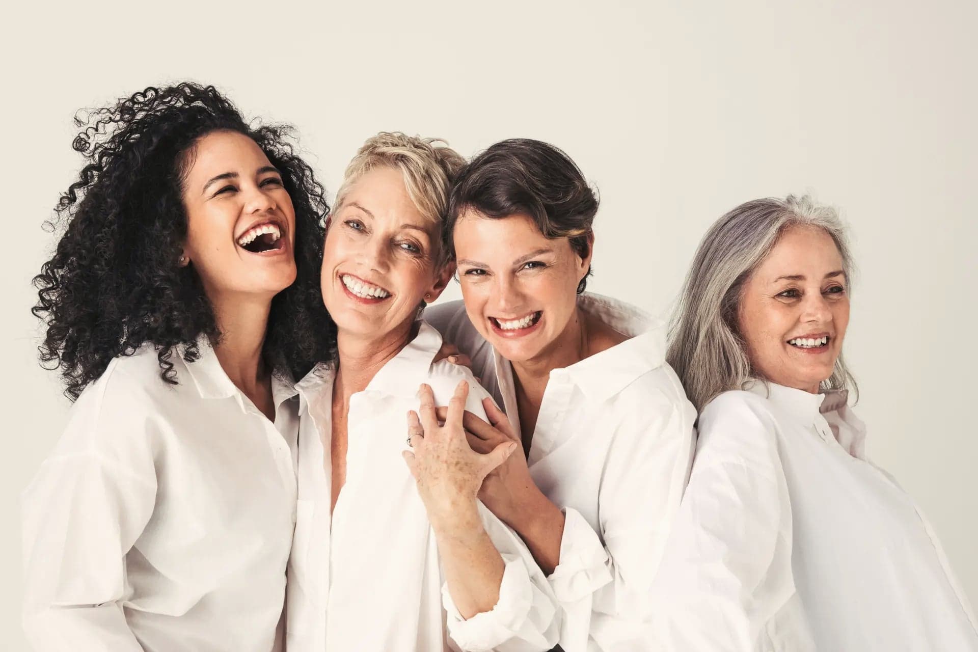 Four women of different ages laughing together in white shirts