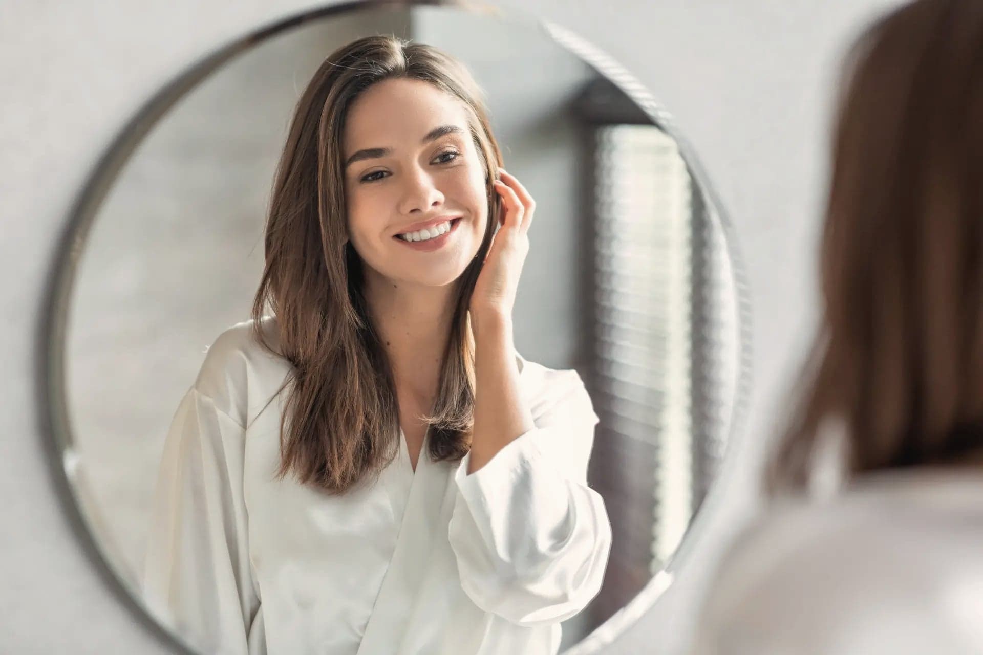 Woman in a white robe looking at her reflection in a round mirror